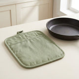 A sage green quilted fabric pot holder with a top loop, displayed on a light cream kitchen table next to a black cast iron skillet.