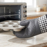 A gray silicone oven mitt with a black-and-white gingham cuff is holding a metal muffin tin over a marble kitchen countertop, with an oven blurred in the background.