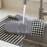 A gray silicone oven mitt with a black and white gingham fabric cuff is rinsed under running water in a stainless steel kitchen sink.