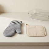 A gray silicone oven mitt with a black-and-white gingham cuff and a beige fabric pot holder with a small zipper pull, arranged on a light countertop beside a clear glass baking dish.