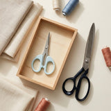 A styled flat-lay of sewing scissors, including a pair with pale blue handles resting in a small wooden tray and another pair with black handles placed beside it, surrounded by beige fabric, thread spools, and a wooden box on a light cream background.