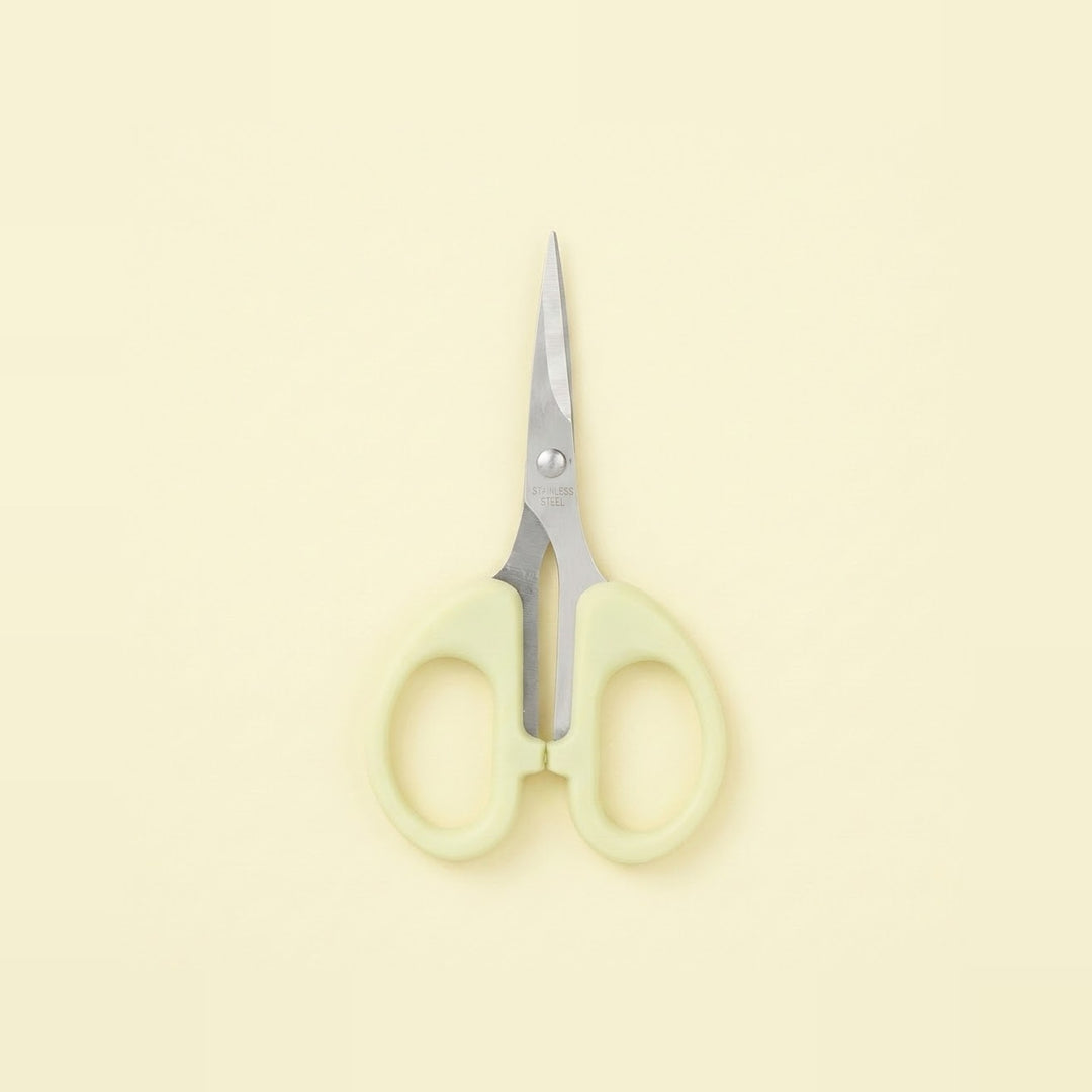 A pair of small scissors with pale yellow-green handles and silver metal blades, centered on a solid pale cream background.