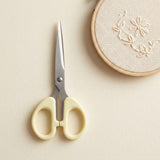 A pair of embroidery scissors with pale yellow handles and silver metal blades, placed on a light beige surface next to a wooden embroidery hoop with floral stitching.