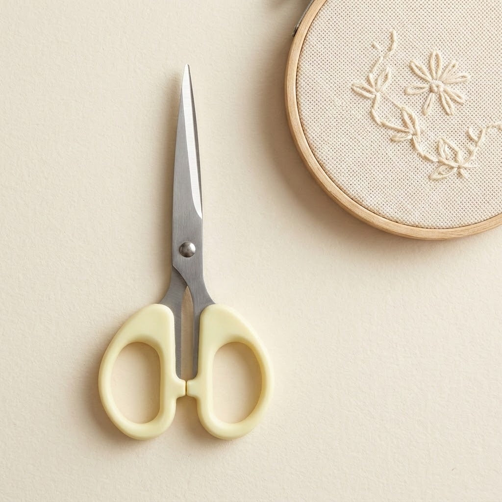 A pair of embroidery scissors with pale yellow handles and silver metal blades, placed on a light beige surface next to a wooden embroidery hoop with floral stitching.