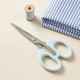 A pair of light blue-handled scissors with silver metal blades, arranged on a beige fabric surface next to a spool of pale blue thread and a folded stack of blue-and-white striped fabric.