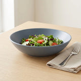A matte slate blue-gray ceramic salad bowl filled with mixed greens, tomatoes, and cucumbers, set on a light wood table next to a beige napkin and silver fork.