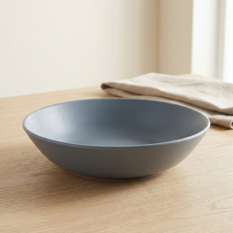 A matte slate blue-gray ceramic bowl resting on a light wood table, with a folded beige cloth napkin blurred in the background.
