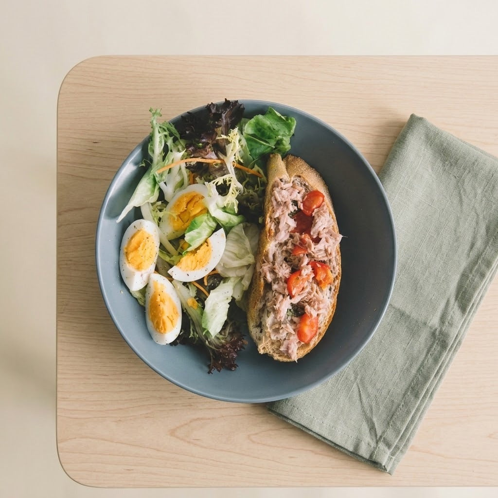 A top-down view of a blue-gray bowl containing a tuna sandwich on bread, mixed greens, cucumber, avocado, and hard-boiled egg slices, placed on a light wood board next to a folded sage green napkin.