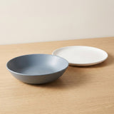 A matte ceramic bowl in a muted blue-gray color and a matching shallow plate in white, displayed on a light wood tabletop against a neutral beige wall.