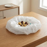 A white crumpled paper bag or dust cover sits on a light wood table in a bright indoor room, with a small pile of mixed snack pieces in the center.