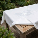 A white waterproof tablecloth draped over a wooden outdoor table, with a glossy water droplet pool demonstrating its water-resistant property. The background shows a blurred garden setting with green foliage and a wooden deck.