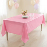 A pink tablecloth draped over a rectangular table in a bright indoor setting, with a small bouquet of flowers, stacked plates, and clear drinking glasses on top. Balloons hang in the background near white curtains.