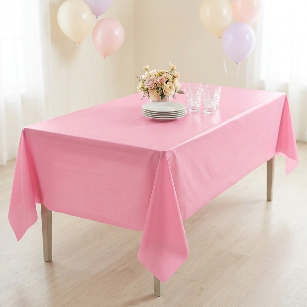 A pink tablecloth draped over a rectangular table in a bright indoor setting, with a small bouquet of flowers, stacked plates, and clear drinking glasses on top. Balloons hang in the background near white curtains.