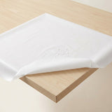 A white tablecloth draped over a light wood table, with a corner folded down to show the fabric's drape and texture.