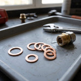 A close-up of plumbing hardware parts arranged on a metal tray, including several copper washers and a brass male connector, with tools and a window in the background.