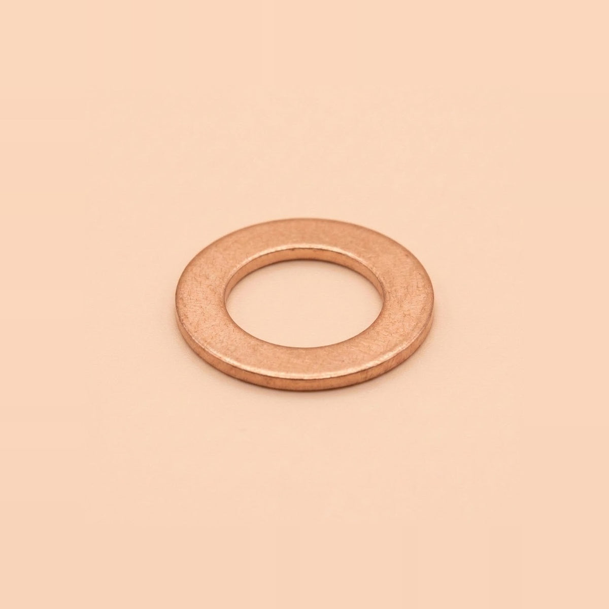 A single flat metal washer with a warm copper or rose-gold metallic finish, centered on a solid, warm peach-beige background.