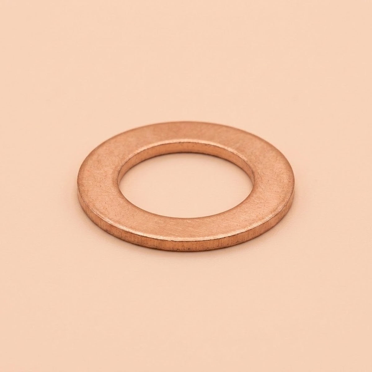 A single flat metal washer with a warm copper or rose-gold metallic finish, centered on a solid, warm beige background.
