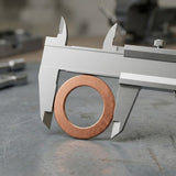 A copper ring or washer is being measured by a silver metal caliper on a gray workbench, with blurred workshop tools in the background.