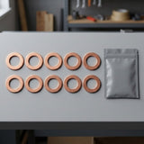 Ten copper-colored circular ring-shaped components arranged in two rows on a gray workbench next to a small gray resealable pouch, with a blurred workshop background.