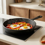 A black cast iron skillet with a handle sits on a modern induction cooktop, cooking two sunny-side-up eggs with sliced red and yellow bell peppers and cherry tomatoes in a bright, contemporary kitchen.