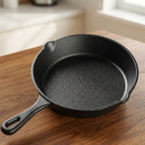 A black cast iron skillet with a handle and small pouring lip, photographed on a wooden tabletop in a softly blurred kitchen setting.