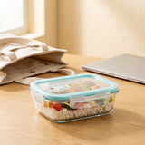 A clear rectangular food storage container with a light blue lid sits on a wooden table, filled with rice and mixed vegetables. A beige tote bag, a closed silver laptop, and a softly lit window are visible in the background.