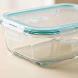 Clear glass food storage container with a translucent lid and a light aqua silicone seal, shown on a beige surface.