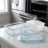 A set of glass and plastic storage containers on a white kitchen countertop, with a stainless steel refrigerator and open oven in the background.