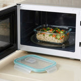 A clear glass rectangular food storage container filled with rice and chopped vegetables sits inside an open white microwave on a glass turntable. The matching clear lid with a light blue silicone seal is placed in front on a beige countertop.