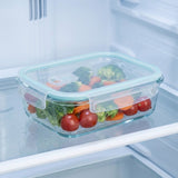 A clear glass food storage container with a light blue silicone seal sits on a refrigerator shelf, filled with broccoli, tomatoes, and carrots.