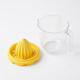 Detached yellow citrus reamer with seeds caught in its slotted tray sits beside an empty clear handled cup on white.
