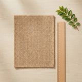 A square beige woven fabric swatch with a coarse burlap texture, displayed on a light wood background next to a wooden ruler and a green leafy sprig.