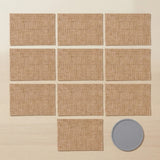 Nine square tan woven placemats arranged in a grid on a light wood surface, with one gray round coaster placed at the bottom right.