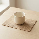 A cream ceramic cup rests on a rectangular beige burlap coaster on a light wooden table beside a window.
