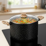 A black speckled pot with a clear glass lid and wooden handles sits on a black stovetop in a softly blurred kitchen setting. Multicolored vegetables are visible inside the pot.