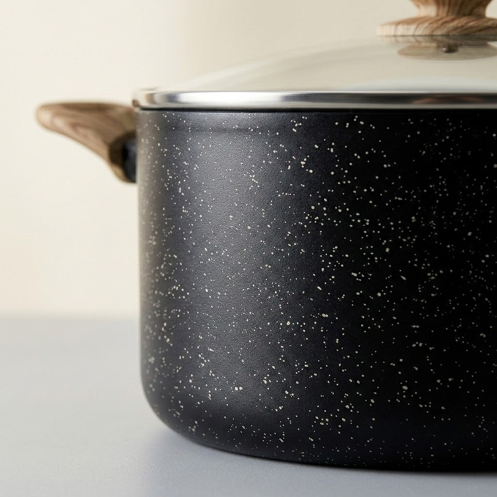 A close-up of a black speckled saucepan with a glass lid and light brown wooden handles, set against a neutral off-white background.