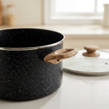 A black speckled saucepan with a light brown wooden handle sits on a light countertop, with a matching glass lid placed behind it in a softly blurred kitchen setting.