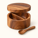 Divided acacia wood salt cellar with removable lid and spoon shown on a cream studio backdrop.