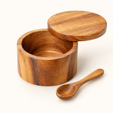 Single bowl acacia wood salt cellar with lid and spoon shown on a cream studio backdrop.