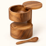 Two-tier acacia wood salt cellar with lids and spoon shown on a cream studio backdrop.