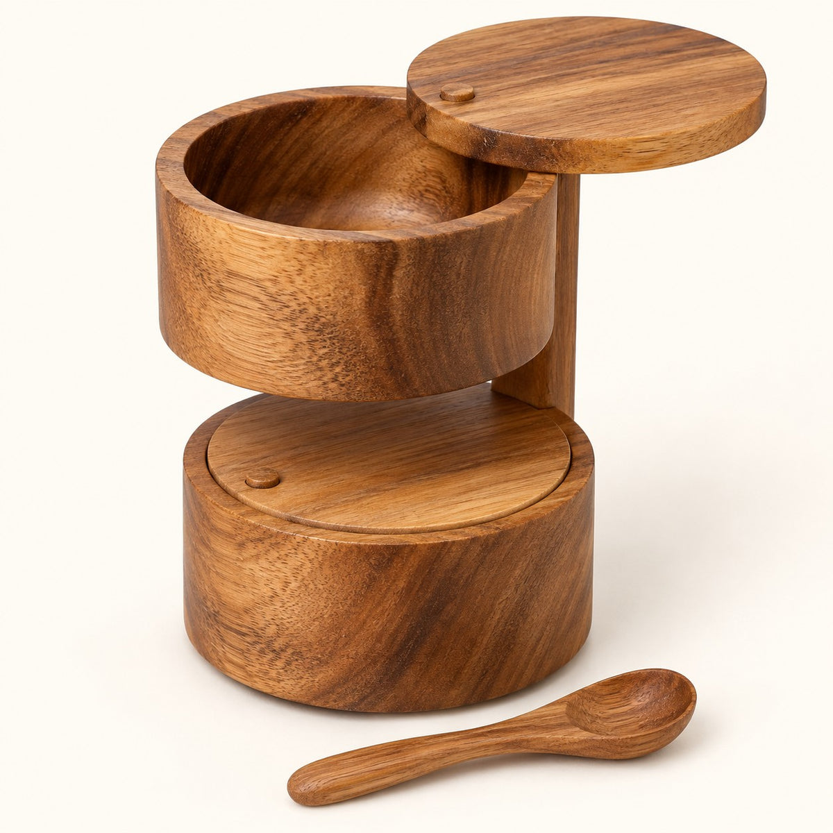Two-tier acacia wood salt cellar with lids and spoon shown on a cream studio backdrop.
