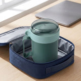 A sage green insulated travel mug with a clear lid and handle, resting inside an open navy blue zippered lunch bag on a light wood desk, with a laptop blurred in the background.