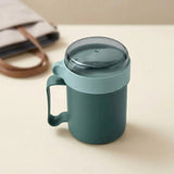 A dark teal insulated mug with a matching handle and a translucent dark gray lid, set on a light beige surface with a soft-focus beige tote bag in the background.