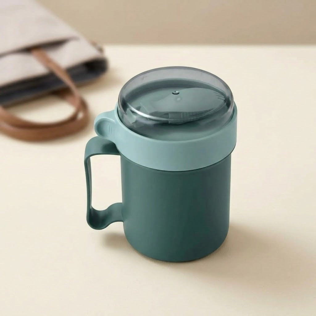 A dark teal insulated mug with a matching handle and a translucent dark gray lid, set on a light beige surface with a soft-focus beige tote bag in the background.