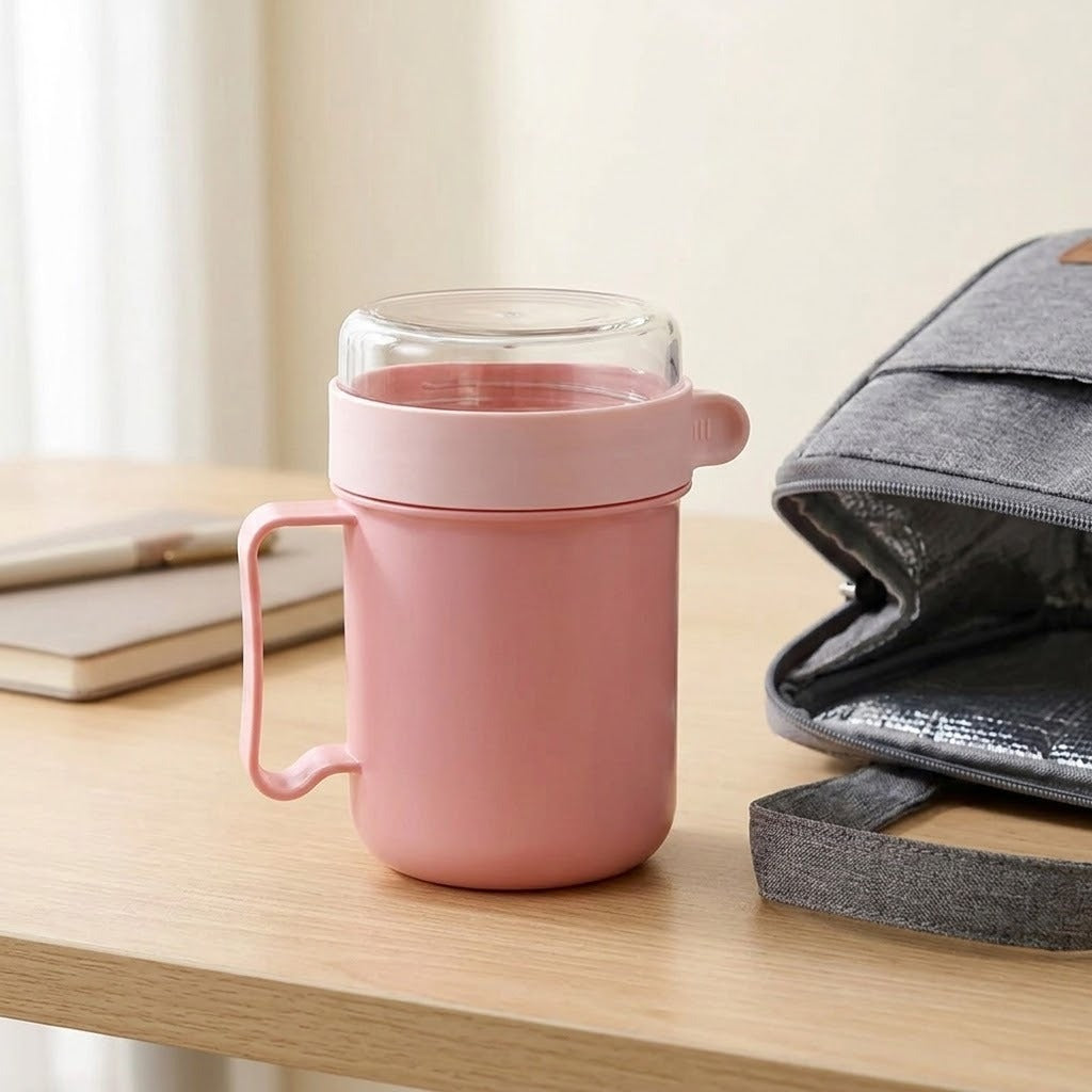 Pink plastic soup mug with clear-topped lid stands on a wooden desk beside notebooks and an open grey insulated bag.