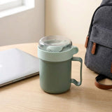 Green plastic soup mug with a folded spoon beneath its clear lid sits on a wooden desk between a laptop and backpack.