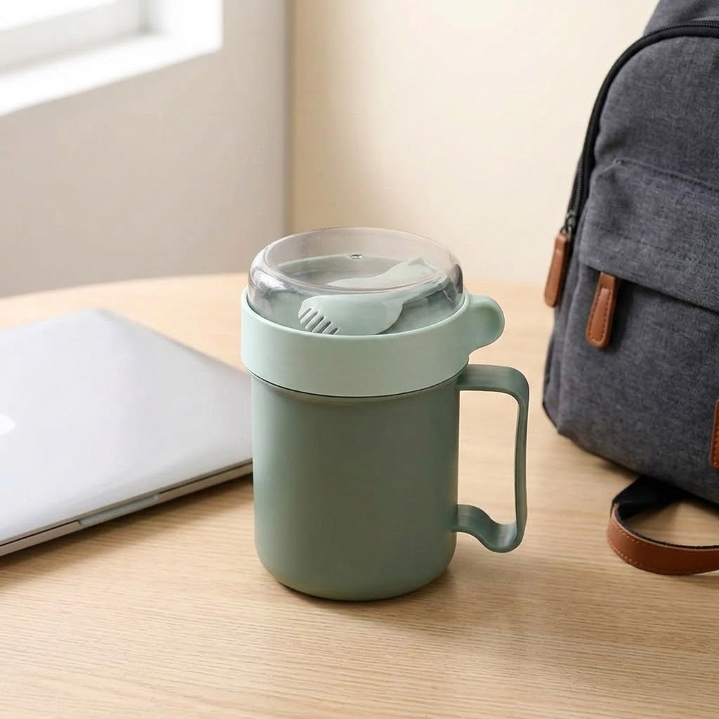 Green plastic soup mug with a folded spoon beneath its clear lid sits on a wooden desk between a laptop and backpack.