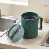 Open green plastic soup mug and detached lid containing a folded spoon sit beside water droplets on a kitchen counter near a sink.