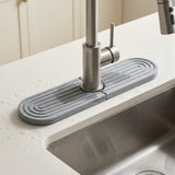 A gray silicone kitchen sink drain guard with concentric oval ridges, shown installed around the base of a stainless steel faucet on a white countertop next to a stainless steel sink.