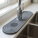 Dark gray grooved diatomite mat surrounds a chrome faucet beside a stainless steel double sink beneath a window.